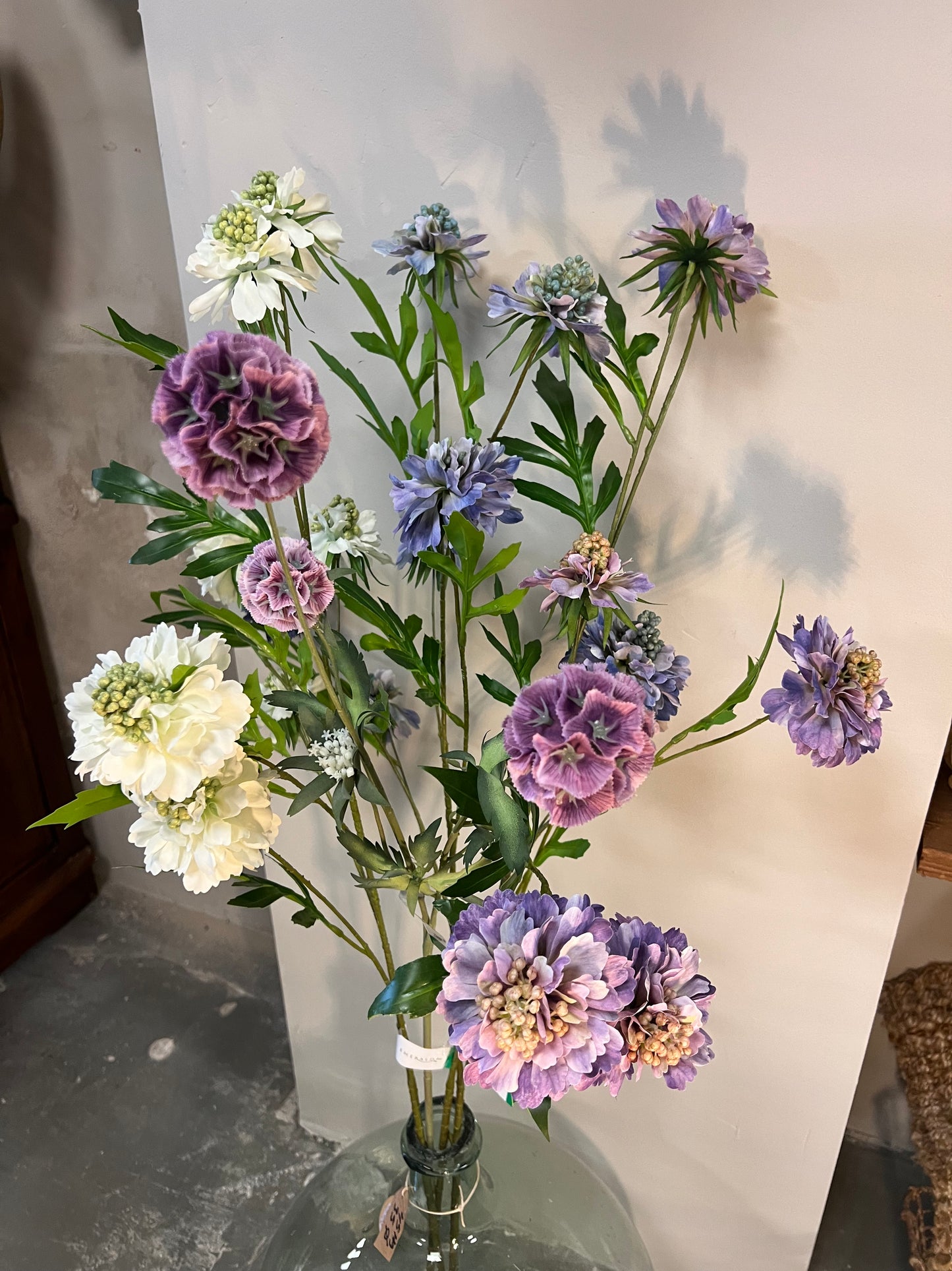 Zijden bloemenarrangement met lila, paarse en witte bloemen in een glazen vaas, tegen een lichte achtergrond