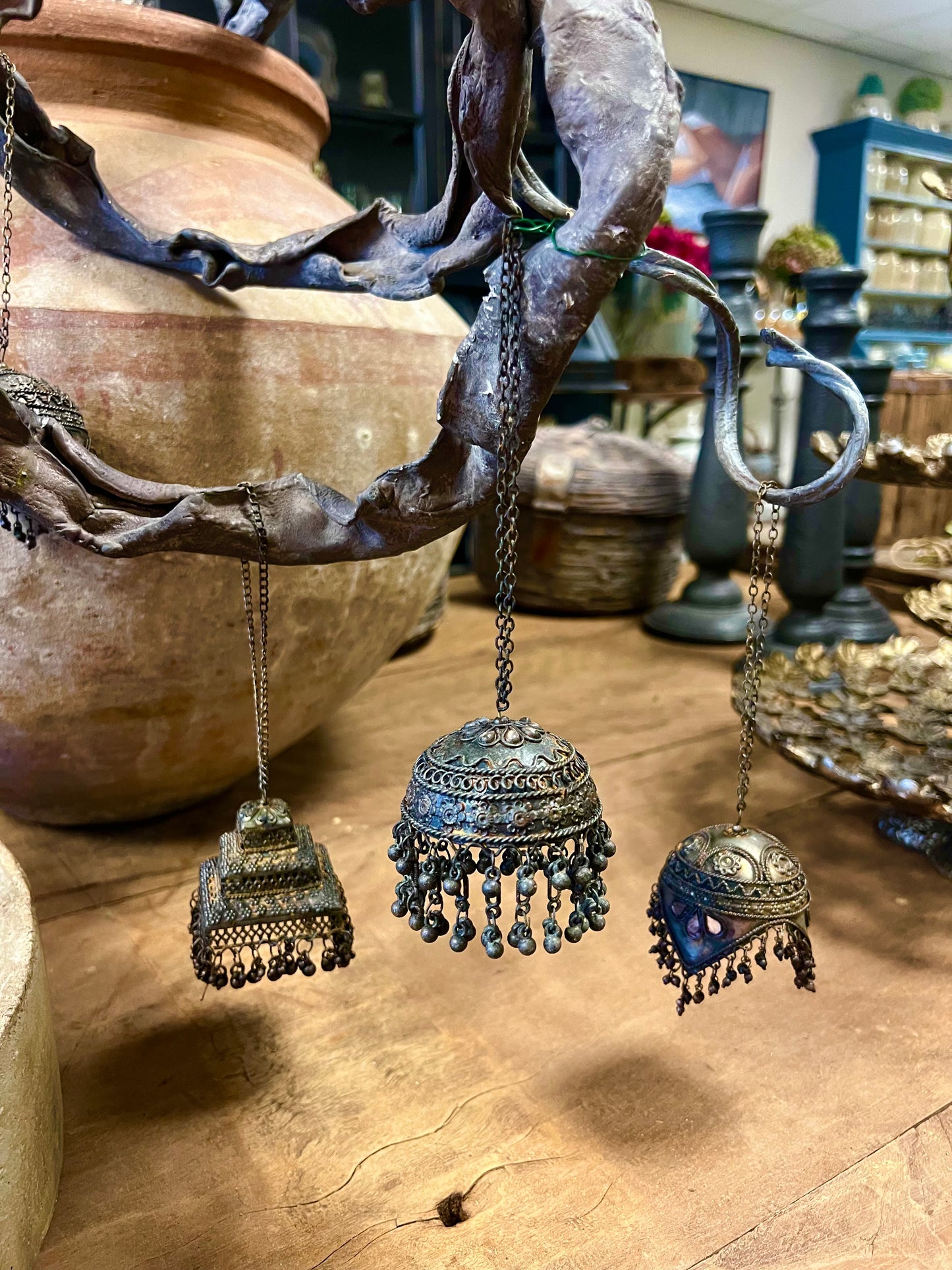 Drie decoratieve metalen ornamenten in de vorm van belletjes, versierd met details, opgehangen aan een tak in een rustieke omgeving.
