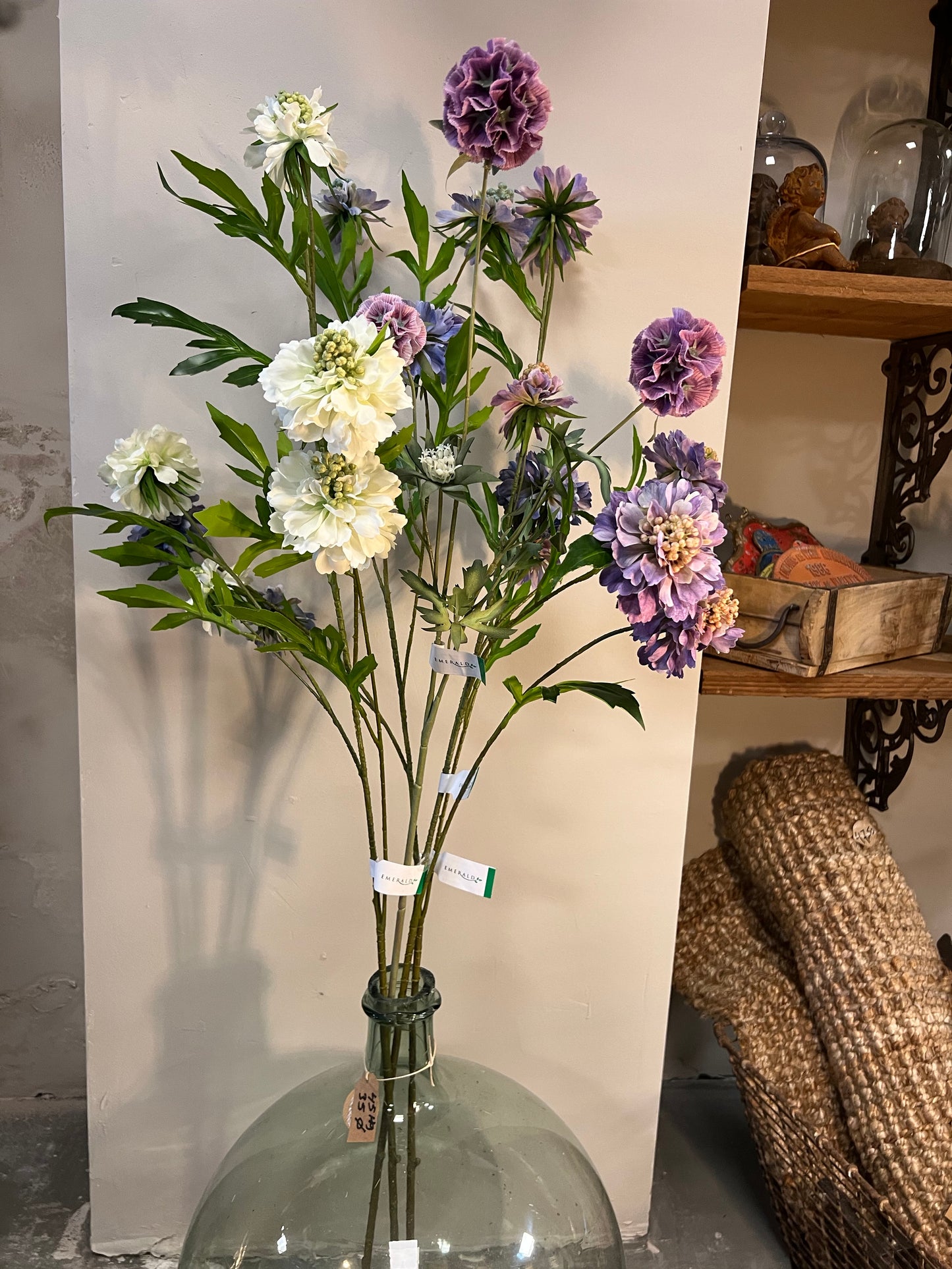 Zijden bloemenarrangement met lila, paarse en witte bloemen in een glazen vaas, tegen een lichte achtergrond
