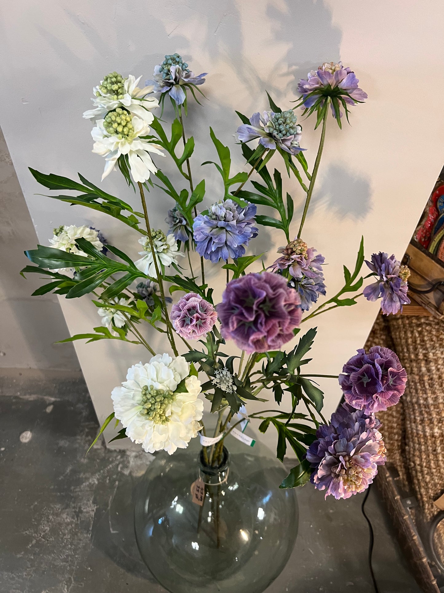 Zijden bloemenarrangement met lila, paarse en witte bloemen in een glazen vaas, tegen een lichte achtergrond