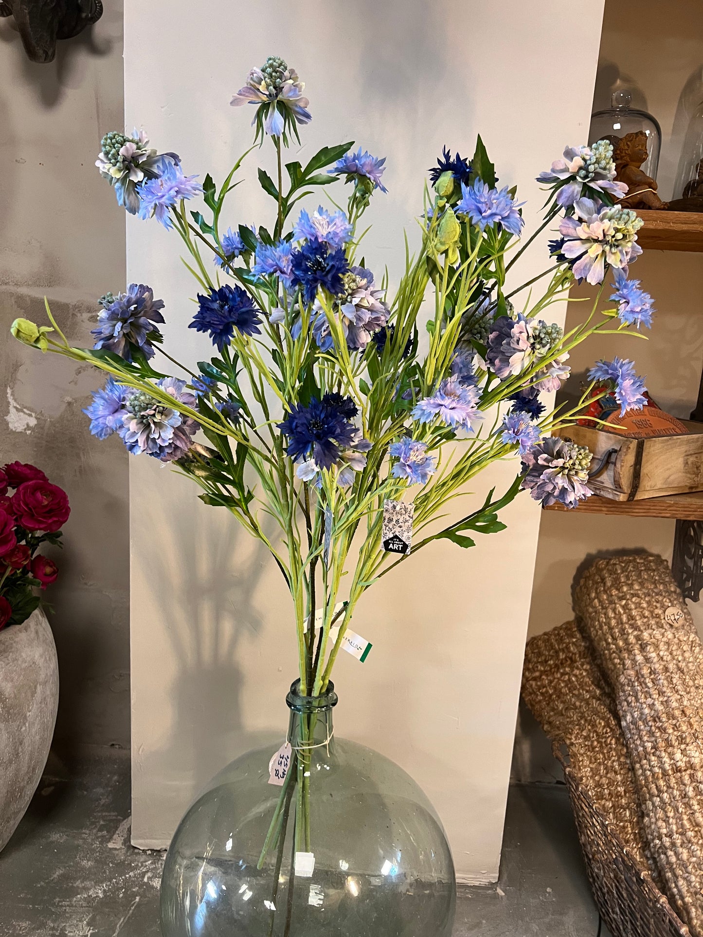 Zijden bloemenarrangement met blauwe en paarse bloemen in een glazen vaas, geplaatst tegen een lichte achtergrond.