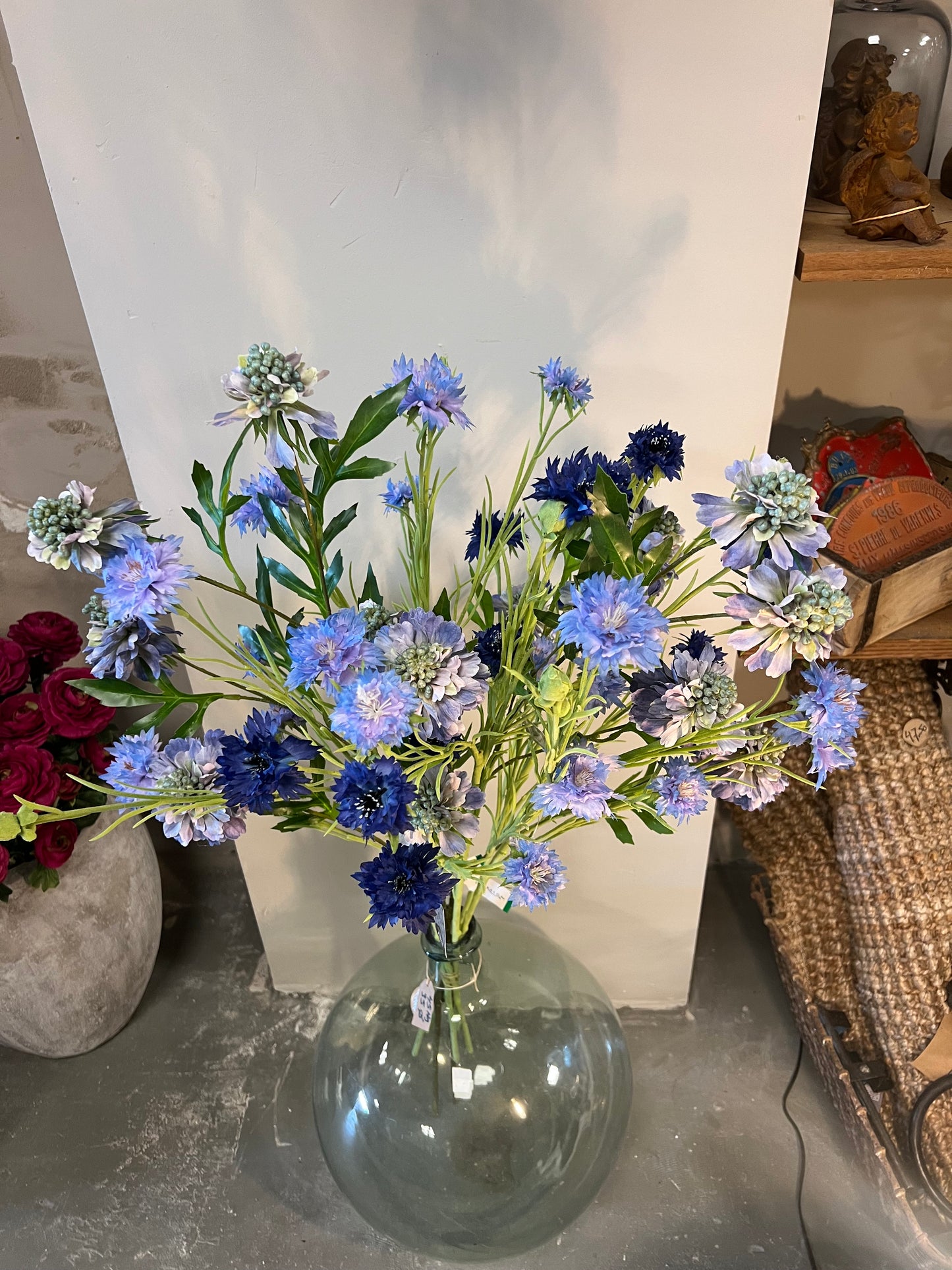 Zijden bloemenarrangement met blauwe en paarse bloemen in een glazen vaas, geplaatst tegen een lichte achtergrond.