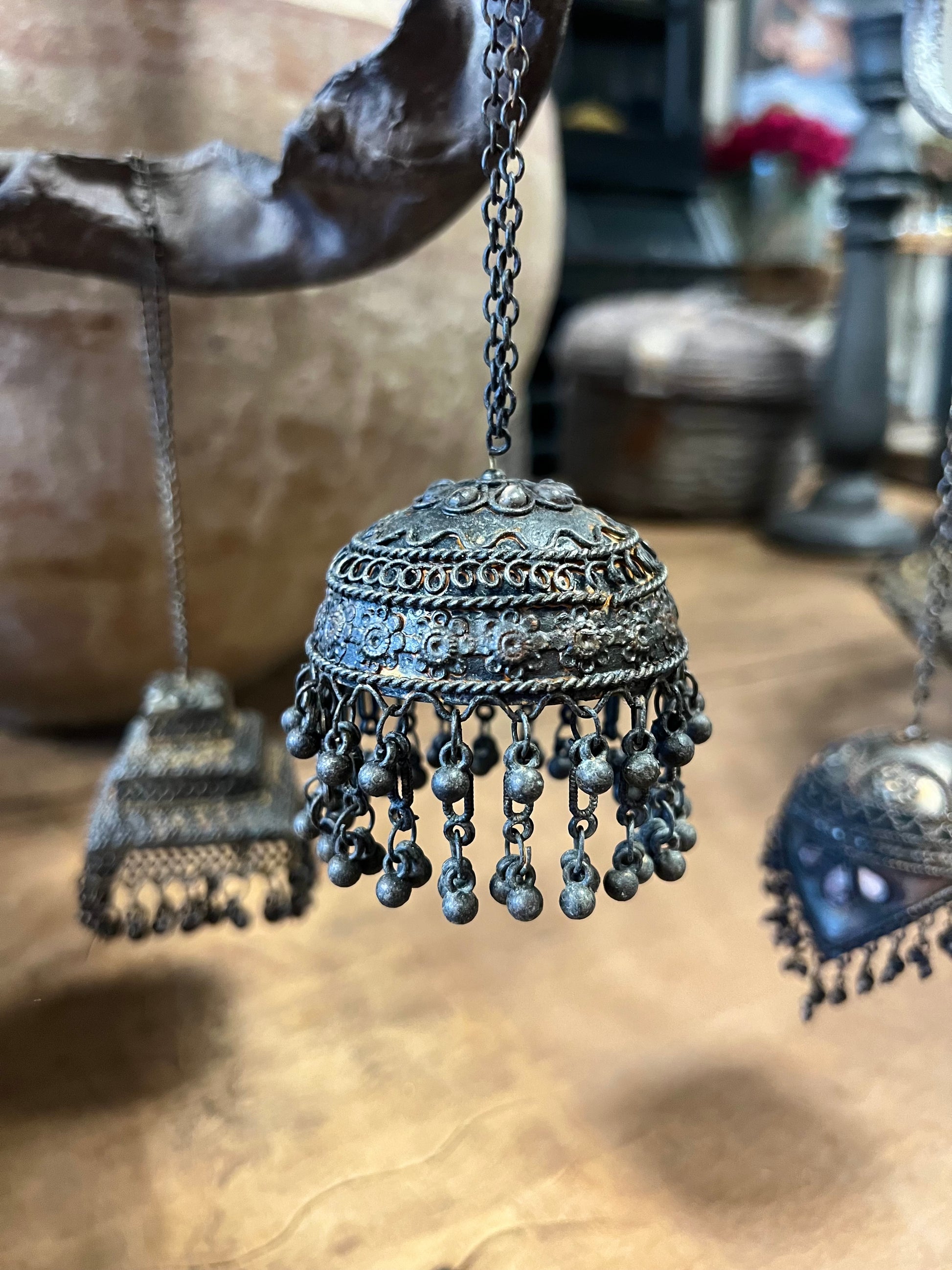 Drie decoratieve metalen ornamenten in de vorm van belletjes, versierd met details, opgehangen aan een tak in een rustieke omgeving.