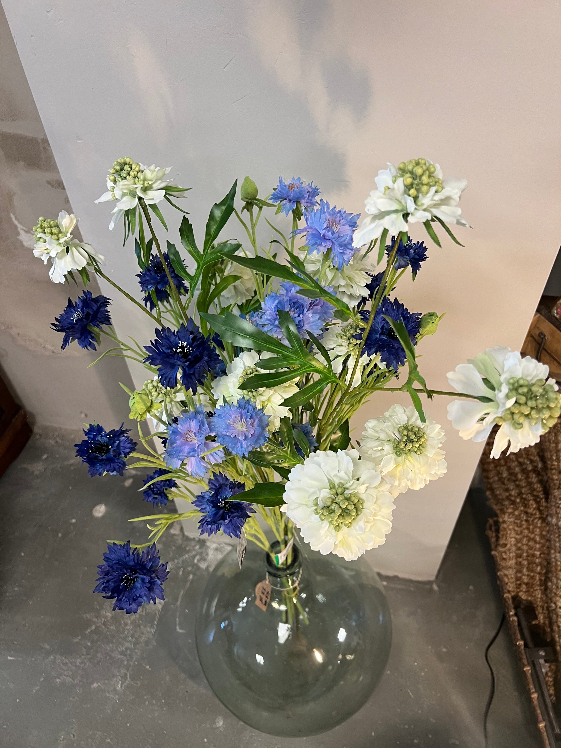 Zijden bloemenarrangement met blauwe en witte bloemen in een rustieke, aardewerken vaas, geplaatst op een houten tafel tegen een lichte achtergrond.
