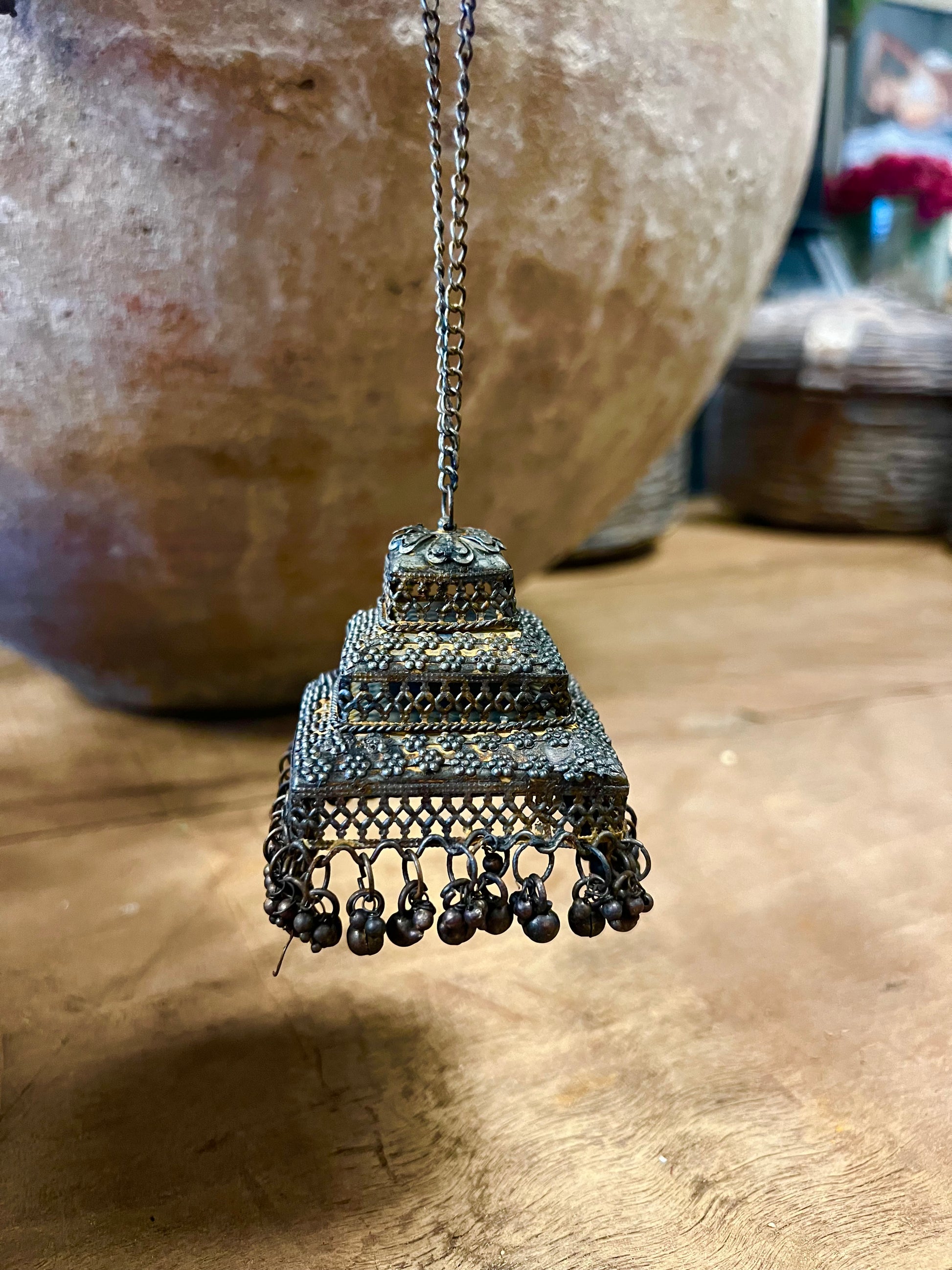 Drie decoratieve metalen ornamenten in de vorm van belletjes, versierd met details, opgehangen aan een tak in een rustieke omgeving.