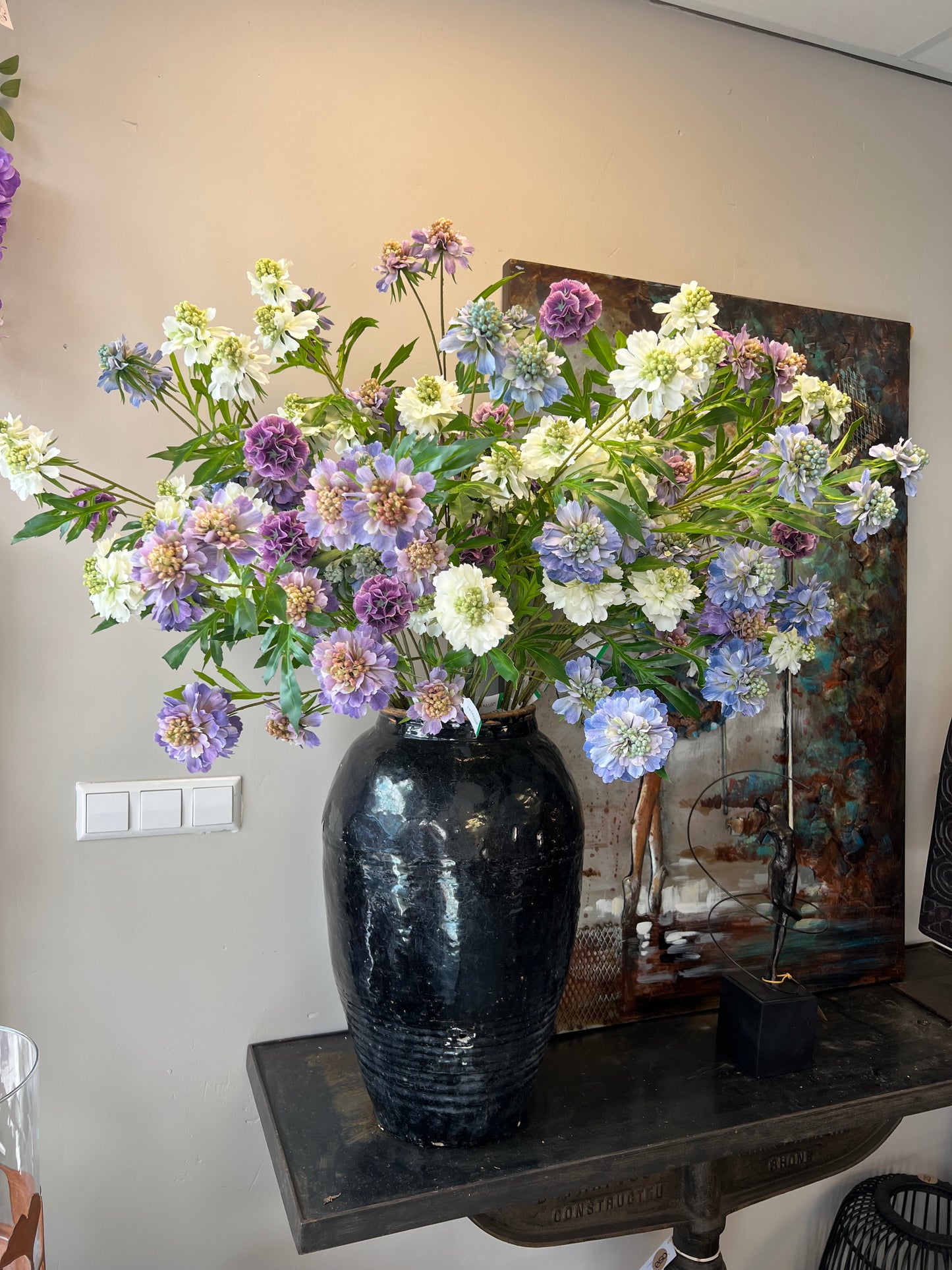 Zijden boeket met lila, paarse en witte bloemen in een zwarte vaas, geplaatst op een tafel.