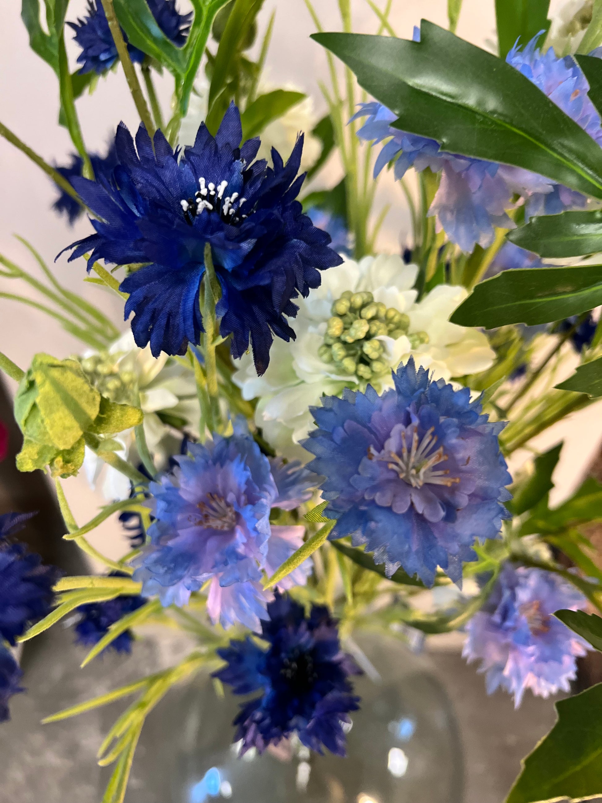 Zijden bloemenarrangement met blauwe en witte bloemen in een rustieke, aardewerken vaas, geplaatst op een houten tafel tegen een lichte achtergrond.