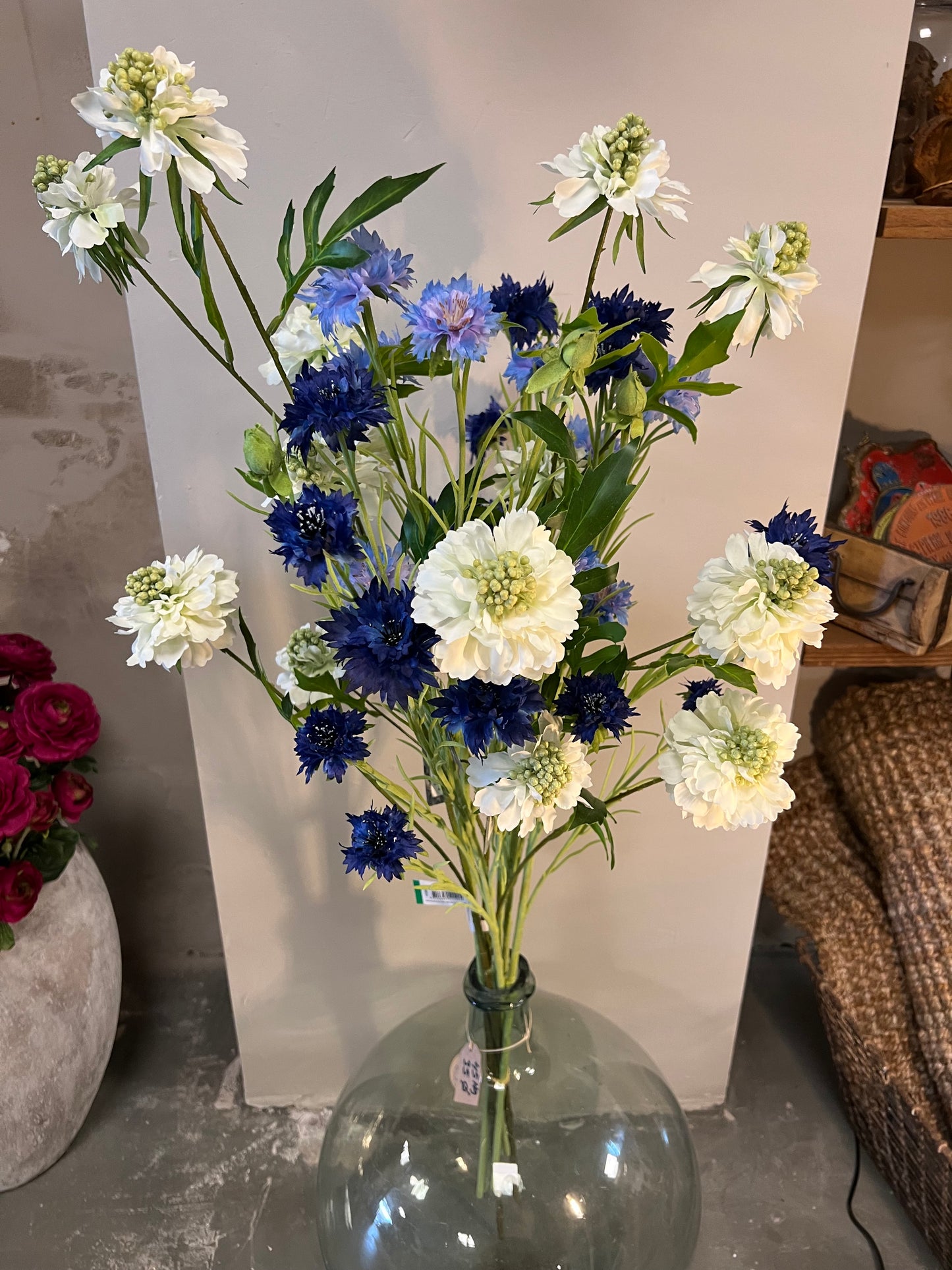 Zijden bloemenarrangement met blauwe en witte bloemen in een rustieke, aardewerken vaas, geplaatst op een houten tafel tegen een lichte achtergrond.