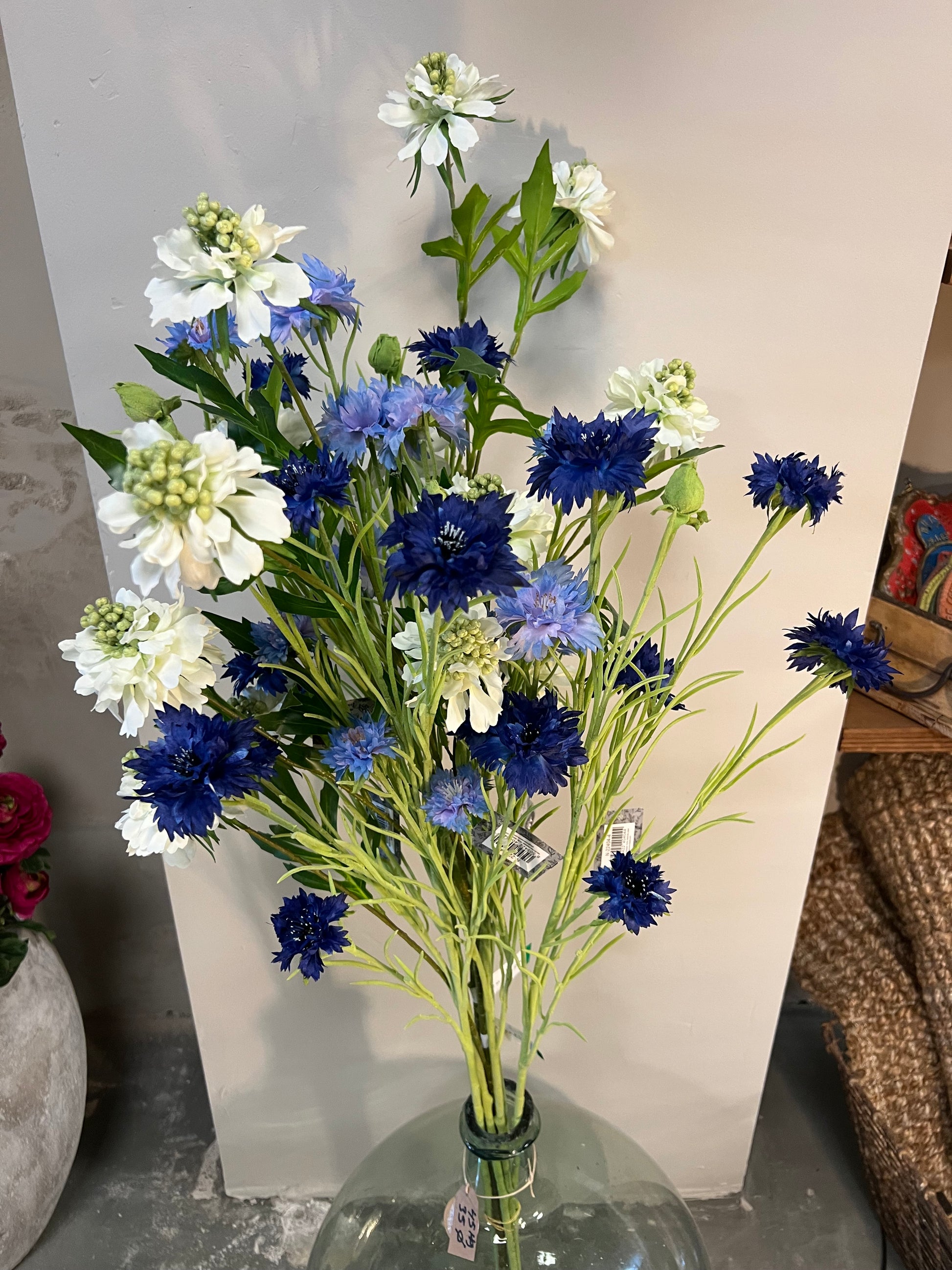 Zijden bloemenarrangement met blauwe en witte bloemen in een rustieke, aardewerken vaas, geplaatst op een houten tafel tegen een lichte achtergrond.