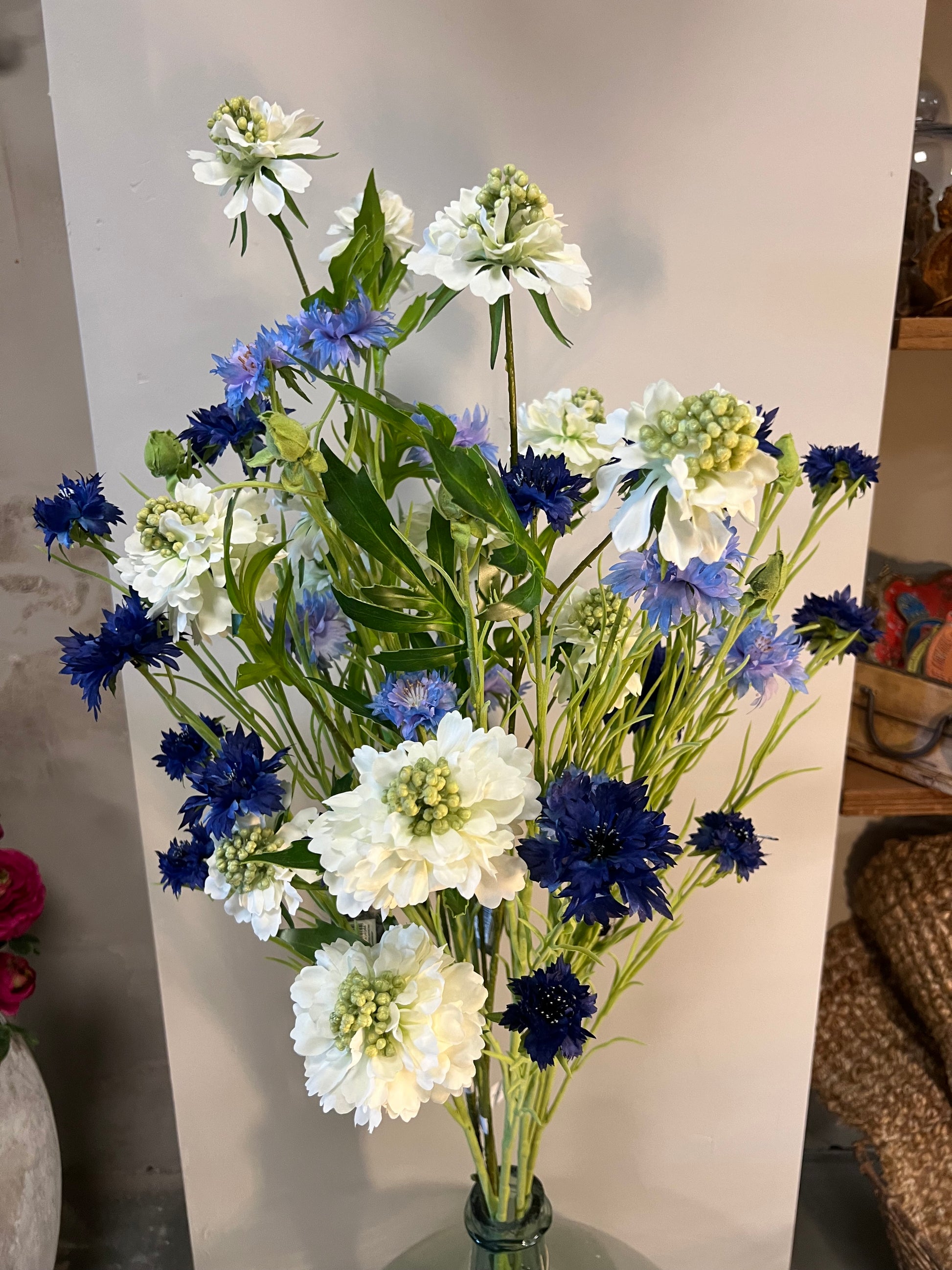 Zijden bloemenarrangement met blauwe en witte bloemen in een rustieke, aardewerken vaas, geplaatst op een houten tafel tegen een lichte achtergrond.