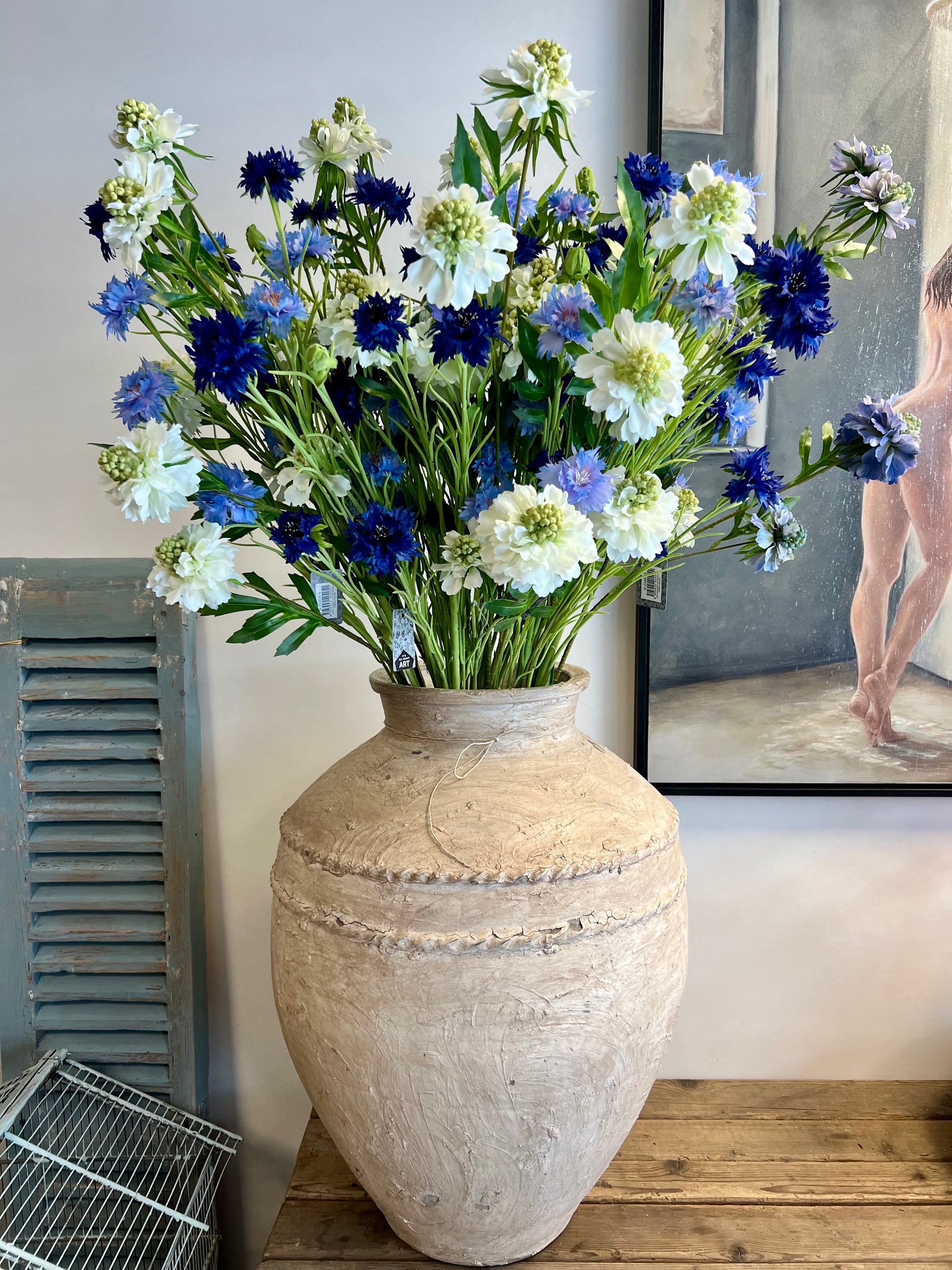 Zijden bloemenarrangement met blauwe en witte bloemen in een rustieke, aardewerken vaas, geplaatst op een houten tafel tegen een lichte achtergrond.