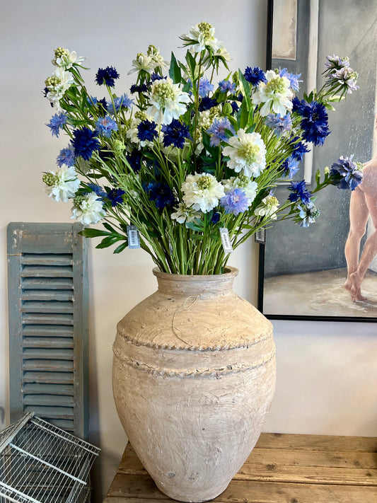 Zijden bloemenarrangement met blauwe en witte bloemen in een rustieke, aardewerken vaas, geplaatst op een houten tafel tegen een lichte achtergrond.