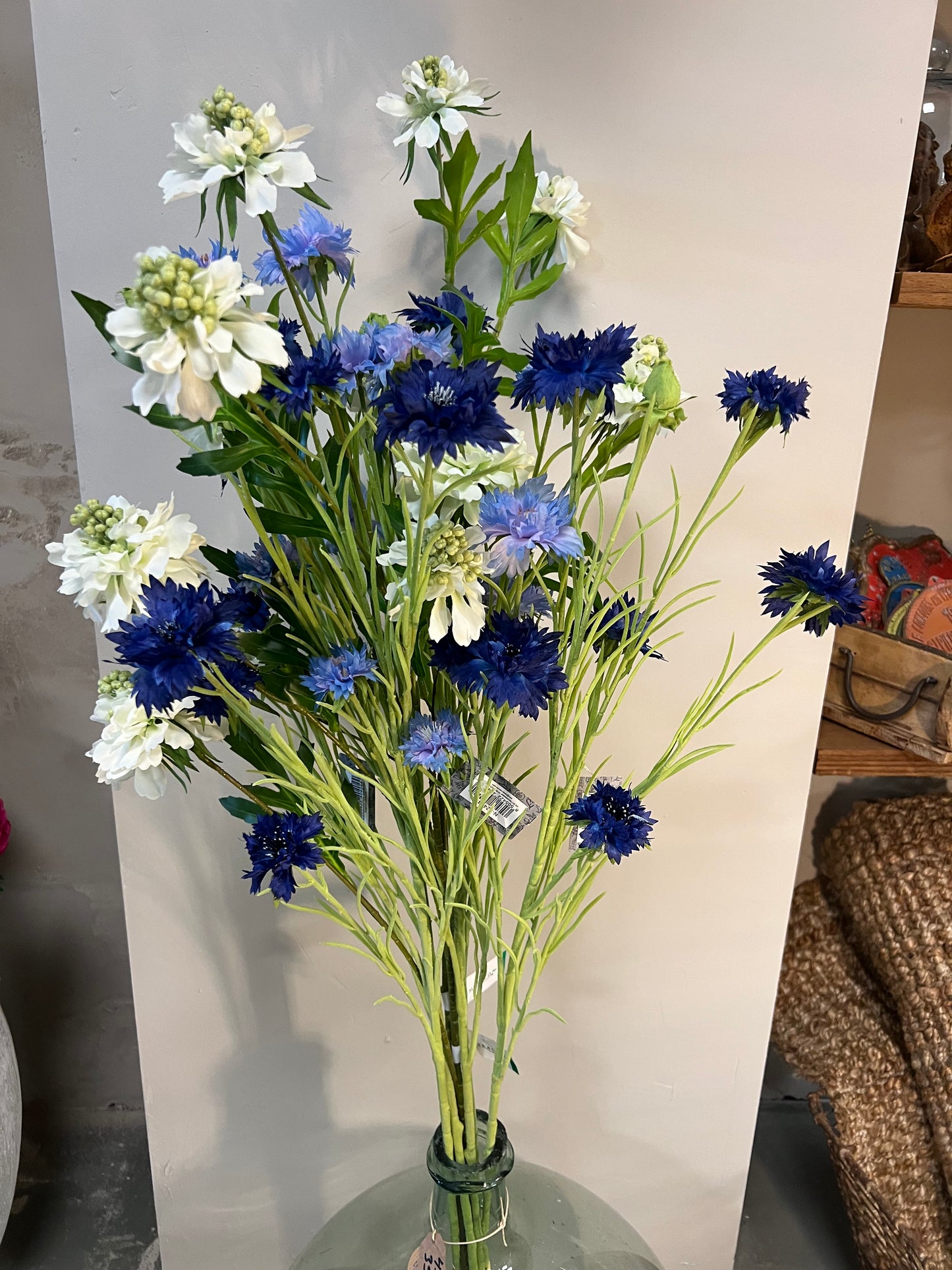 Zijden bloemenarrangement met blauwe en witte bloemen in een rustieke, aardewerken vaas, geplaatst op een houten tafel tegen een lichte achtergrond.