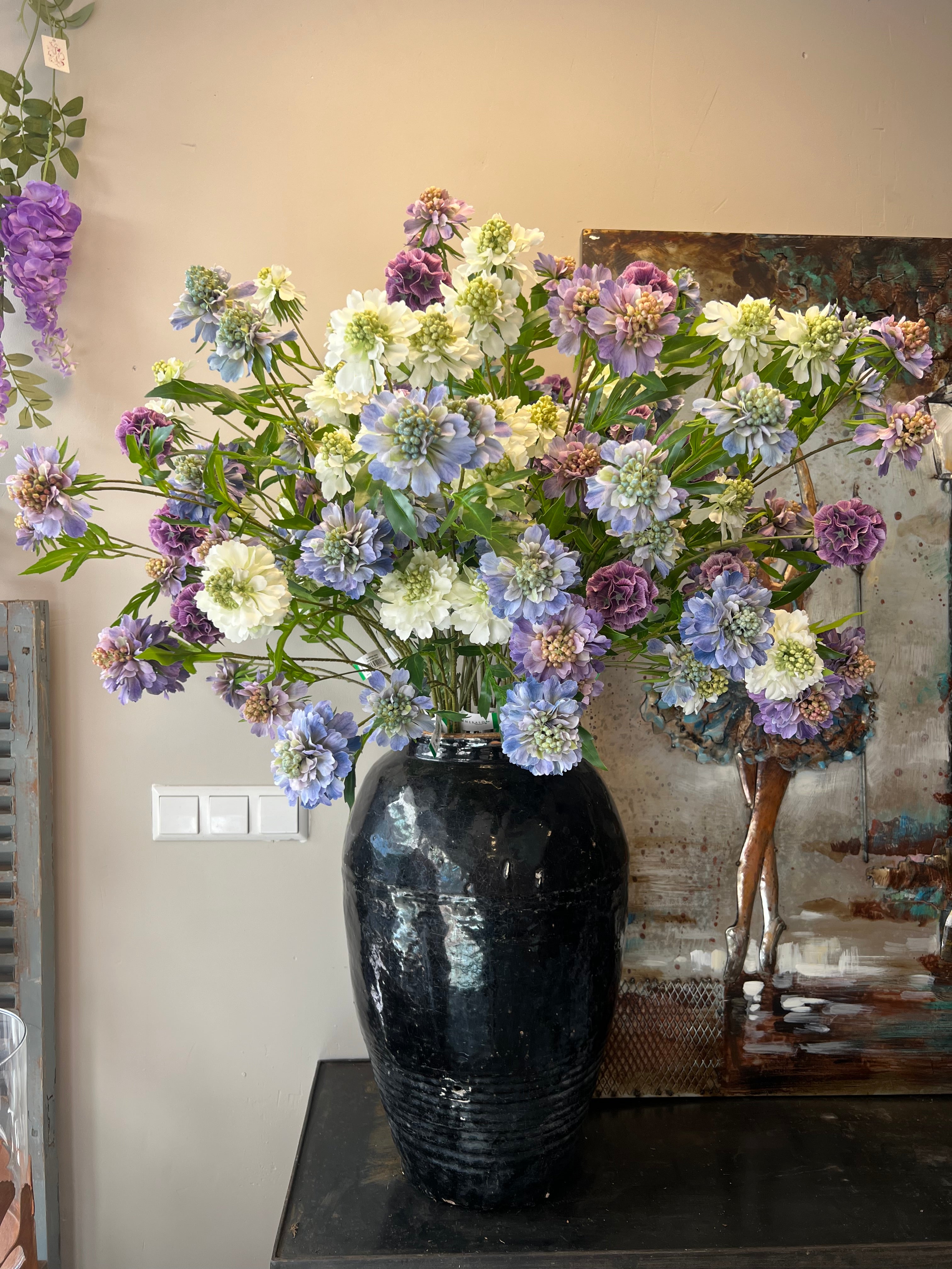 Zijden boeket met lila, paarse en witte bloemen in een zwarte vaas, geplaatst op een tafel.