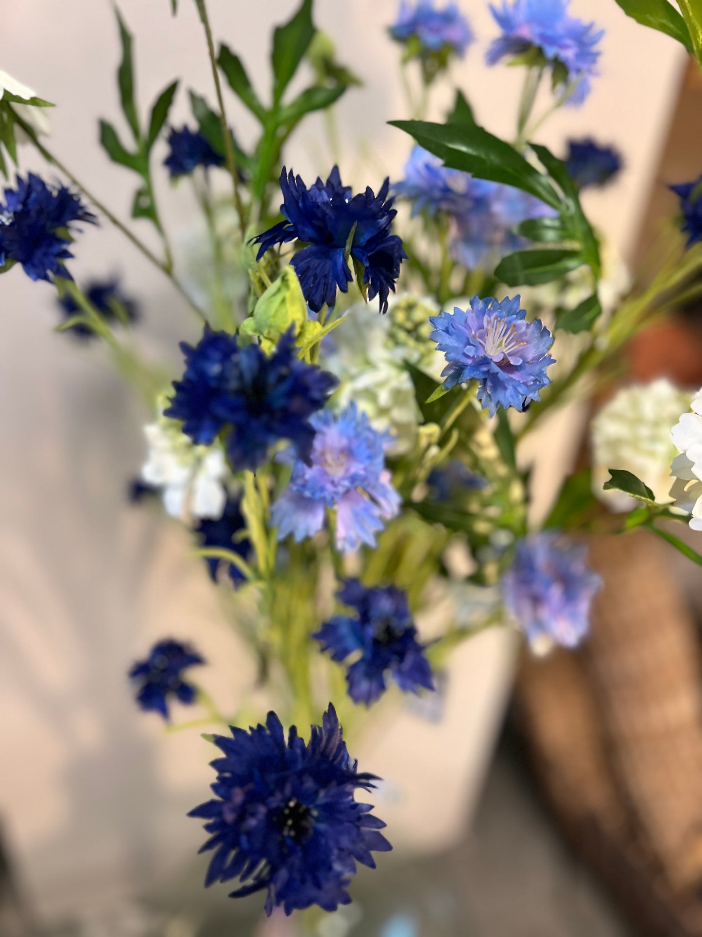 Zijden bloemenarrangement met blauwe en witte bloemen in een rustieke, aardewerken vaas, geplaatst op een houten tafel tegen een lichte achtergrond.